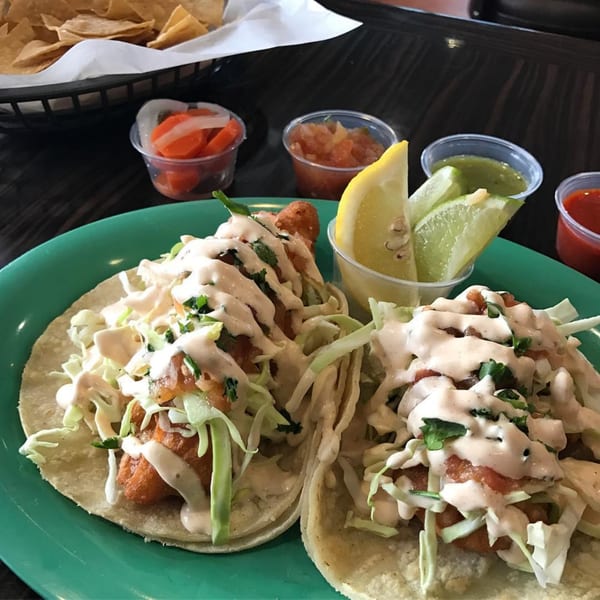 Two Chilitos Mexican Grill fish tacos with cabbage, sauce, and sides of salsa, lime, and chips.